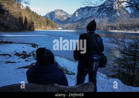 A couple in winter at a mountain lake in the Alps. The woman sits on a bench and rests. The man stands and looks at the mountains. Stock Photo