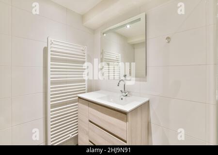 Ceramic sink on the wooden cupboard and a mirror on the wall near to the heating system in a bright white bathroom Stock Photo