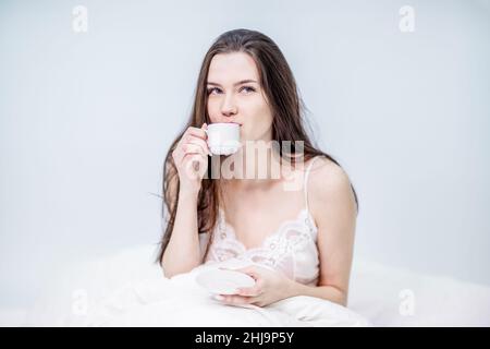 A cute woman is sitting in bed and drinking aromatic coffee. Tasty breakfast. The brunette is holding a cup with a hot drink. White bedroom. Good morn Stock Photo