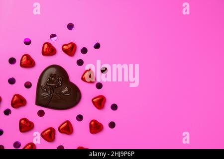 Chocolate heart and lots of shiny candy in the shape of hearts wrapped in foil on a pink background. Beautiful background for a postcard. banner for t Stock Photo