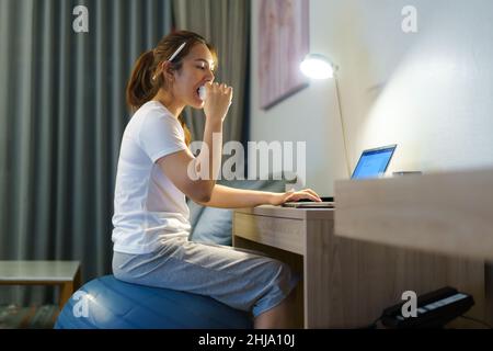 Asian woman relaxing on yoga ball and eating sandwich and chatting online with team of colleagues on laptop while work from home at home at night. Stock Photo