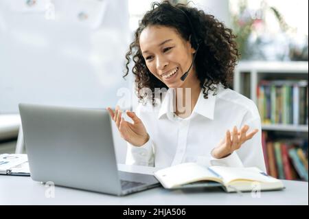 Distant support service, business meeting. African American positive young woman with headset, sit in modern office, talking with colleagues or having online consultation with clients by video call Stock Photo