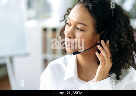 Close-up of black curly haired young woman with headset, female african american support service worker, call center operator, in modern office, looking away, smiling Stock Photo