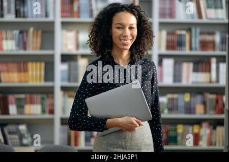 Portrait of lovely pleasant positive African American young woman, manager, freelancer or teacher with a laptop, standing in a modern office or library, looking at the camera, smiling friendly Stock Photo