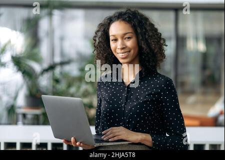 Portrait of curly attractive positive african american pleasant young business woman, freelancer, real estate agent or manager, holding open laptop in office, looking at the camera, smiling friendly Stock Photo