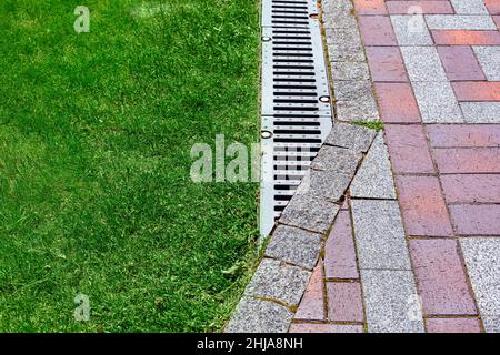 drainage grate bolted to storm drain at corner of pedestrian pavement walk way made of stone brick tiles gray and red pattern in backyard with copy sp Stock Photo
