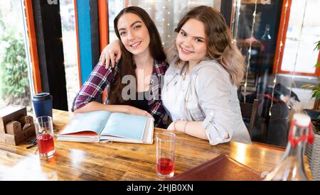 Two girlfriends at cafe smiling looking at camera taking a break in a studying taking photos. Student girls study together while having a lunch break sitting at cafe.  Stock Photo