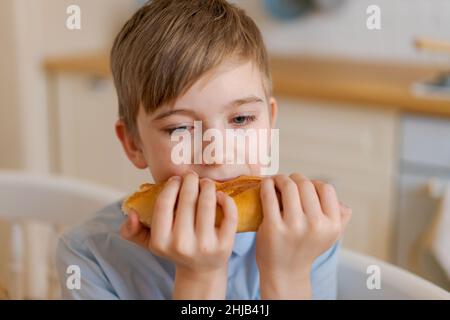 Happy handsome young teenage boy holding and eating freshly baked bread. A hungry boy is biting a big bread, in the kitchen at home. A child with handmade wheat flour bread looks into the camera. Stock Photo
