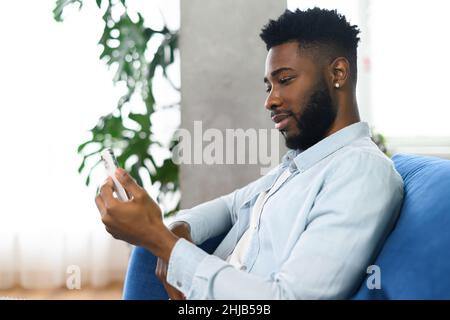 Portrait of cheerful young black guy sending message on mobile phone while sitting at the sofa. Happy multiracial man using smartphone, enjoying chatting online, messaging in social networks Stock Photo