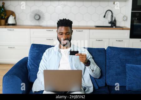 Happy multiracial guy holding credit card and paying online, using laptop computer for shopping online, transferring money, Man sits on the couch and input banking card data for purchasing Stock Photo