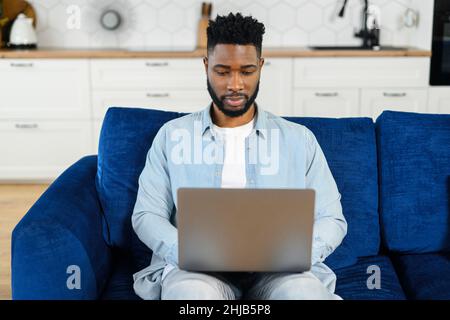 Cheerful man wearing casual clothes sitting on the sofa and using laptop at home. Inspired glad ethnic male entrepreneur typing on keyboard and looks at the screen with pleasure smile Stock Photo