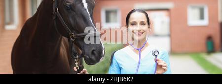 Portrait of young smiling female veterinarian holding steepler while standing next to horse Stock Photo