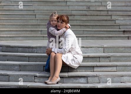 Child hugging mother with love on stairs and looking aside Stock Photo