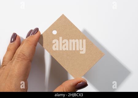 Fingers holding cardboard blank rectangular label tag with small hole in upper part for clothes in center with shadows falling on white background Stock Photo