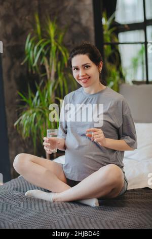 Supplements during pregnancy. Young happy pregnant woman taking prenatal vitamins, holding glass of water and pill while sitting on bed at home. Expec Stock Photo