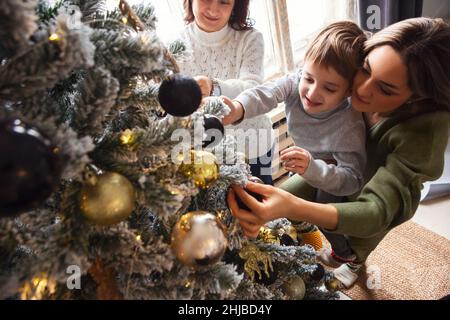 Happy family decorating Christmas tree together at home. Cute little boy hanging toy balls while preparing house for winter holidays with mother and g Stock Photo