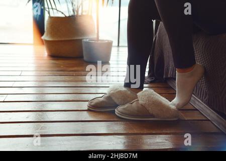 Cropped shot of unrecognizable woman putting on soft plush warm slippers, sitting on sofa in morning time at cozy home. Female wearing fluffy fur hous Stock Photo