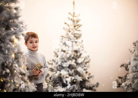 Christmas miracle. Cute happy little boy with teddy bear surrounded with snow-covered artificial xmas trees decorated with yellow twinkle lights, sele Stock Photo