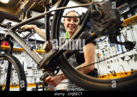From below happy woman smiling and looking at camera while spinning wheel of bicycle during work in repair shop. Stock Photo