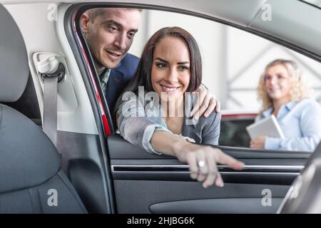 Young couple checks the features of new car interior while the sales manager stands aside in teh car dealership. Stock Photo