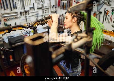 High angle side view of informal female master with dreadlocks fixing bike handlebar in repair service while using screwdriver Stock Photo