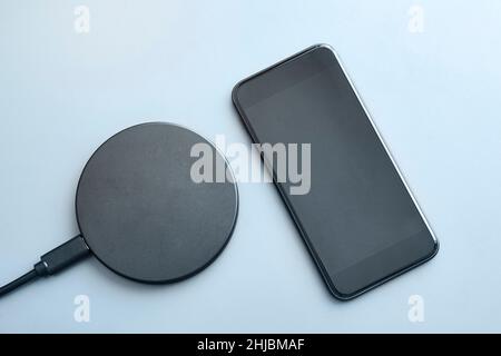 High angle view of modern smartphone with dimmed display next to round wireless charger on light blue background. Modern technology, wireless device a Stock Photo