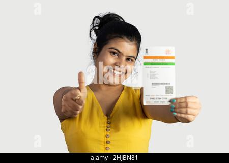 An Indian young woman smiling and showing thumbs up with Aadhaar card in hand on white background Stock Photo