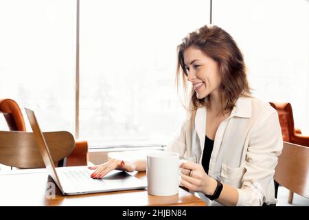 Happy young beautiful business female sitting at the desk, using laptop and hold a cup of coffee. Joyful attractive stylish woman employee is working Stock Photo