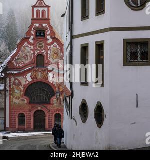 Four tourists walk one after the other along an ascending street in the old town of Füssen. Behind them the Holy Spirit Hospital Church. Stock Photo