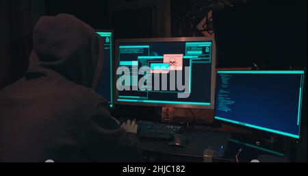 Hacker in a hood sits in front of computer screens and hacks databases Stock Photo