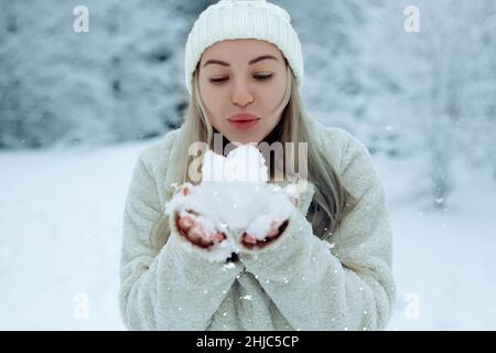 Beautiful girl in fashionable winter clothes blows snow from the palms of  her hands. Joy to the first snow .Young blonde in a white hat in a snowy  forest Stock Photo by ©