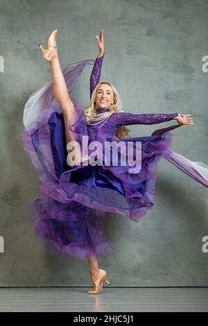 Blonde Female Ballroom dancer in flowing dance movement in purple ballroom dress, against a grey backdrop in the photo studio Stock Photo