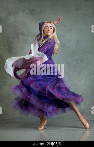 Blonde Female Ballroom dancer in flowing dance movement in purple ballroom dress, against a grey backdrop in the photo studio Stock Photo