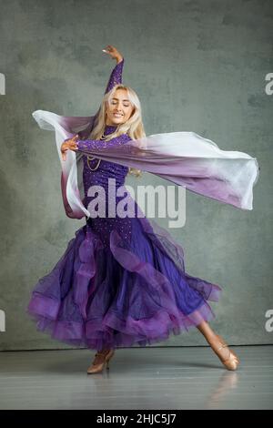 Blonde Female Ballroom dancer in flowing dance movement in purple ballroom dress, against a grey backdrop in the photo studio Stock Photo