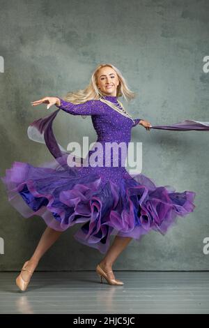 Blonde Female Ballroom dancer in flowing dance movement in purple ballroom dress, against a grey backdrop in the photo studio Stock Photo