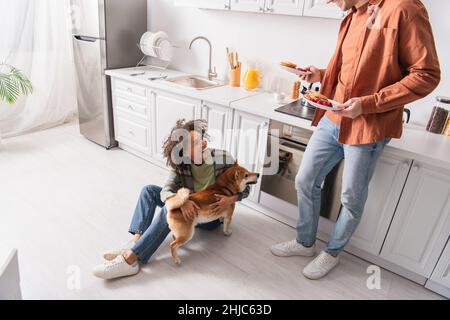 man holding breakfast near cheerful african american girlfriend having fun with shiba inu dog on kitchen floor Stock Photo