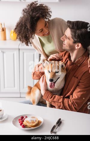 shiba inu dog sticking out tongue near pancakes and happy interracial couple Stock Photo