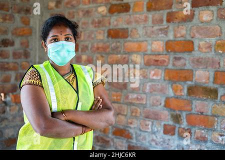 Confident Woman construction employee with medical face mask and safety jacket standing by looking camera with crossed arms at workplace - concept of Stock Photo