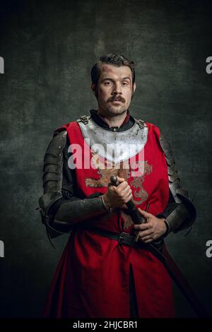 Vintage style portrait of brutal seriuos man, medieval warrior or knight with dirty wounded face holding sword isolated over dark background Stock Photo