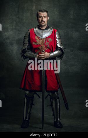 Portrait of brutal seriuos man, medieval warrior or knight with dirty wounded face holding sword isolated over dark background. Comparison of eras Stock Photo