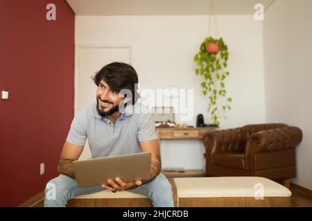 Young business man working at home from his living room chair with laptop on his lap. Home office concept. Gray notebook for working. Home office conc Stock Photo