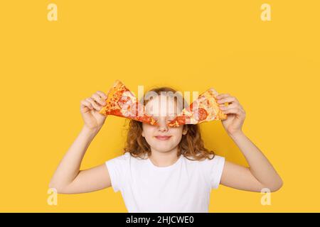 Hungry girl dressed in white t-shirt, holds two slices of pizza and closed eyes from pleasure, photo in yellow background. Stock Photo