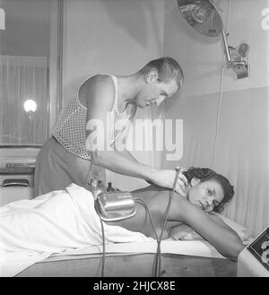 Beauty care in the 1950s. A young woman is having a massage by a man handling the treatment. He is wearing the very special kind of sleeveless shirt with the fabric knitted as a net, mainly to be used under your shirt. An odd looking machine is being used and its purpose is unknown but assumably it was the 1950s topline of treating aching muscles and ease pain. Sweden 1951.  Ref Kristoffersson BE104-9 Stock Photo