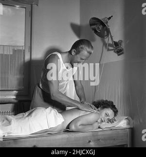Beauty care in the 1950s. A young woman is having a massage by a man handling the treatment. He is wearing the very special kind of sleeveless shirt with the fabric knitted as a net, mainly to be used under your shirt. An odd looking machine is being used and its purpose is unknown but assumably it was the 1950s topline of treating aching muscles and ease pain. Sweden 1951.  Ref Kristoffersson BE104-8 Stock Photo