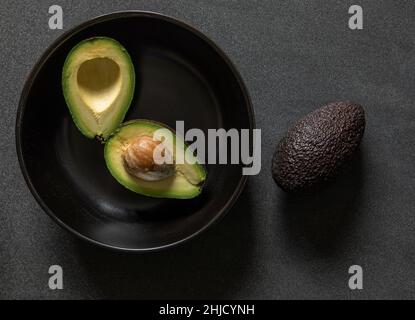 two halves of an avocado one half with the stone in a black bowl another avocado outside the bowl on a dark black textured background from above Stock Photo