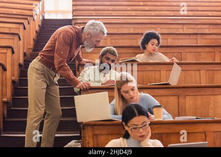 Mature teacher standing near multiethnic students writing on notebooks near laptops in university Stock Photo