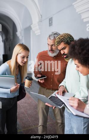 Teacher holding smartphone and talking to multiethnic students with laptops and notebooks in university Stock Photo