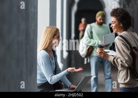 Smiling multiethnic students with coffee to go and laptop talking in corridor of university Stock Photo