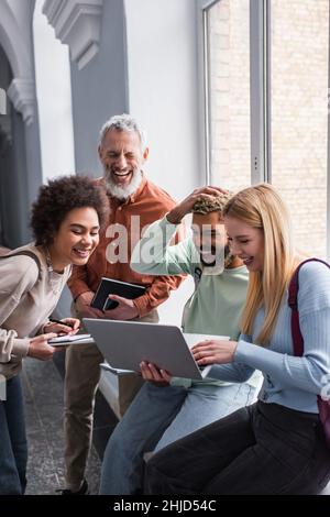 Positive multiethnic students using laptop near teacher with notebook in university hall Stock Photo