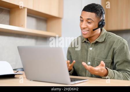 Concentrated young guy in smart casual wear is using headset and laptop for online communication, supporting, selling. Black guy sitting at the office desk Stock Photo